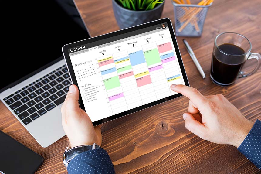 A person holding a tablet with a digital calendar open, reviewing a schedule at a desk with a laptop, coffee, and stationery.