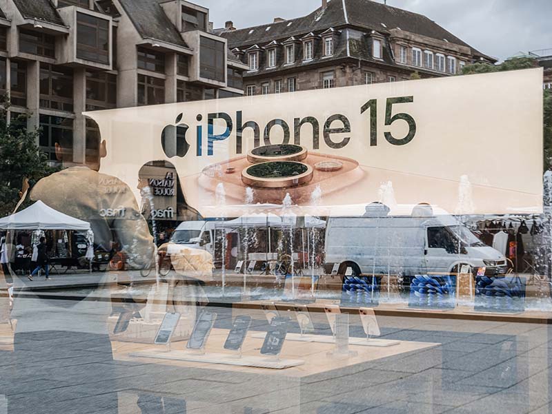 Photo seen through the store window of people looking at various iPhones on display inside an Apple store with an iPhone 15 banner.]