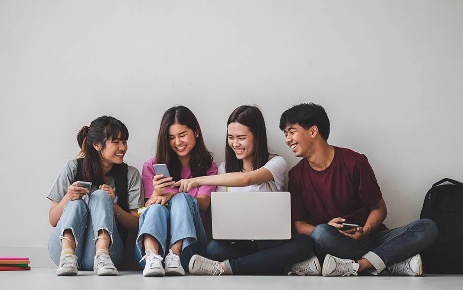 Smiling students sit comfortably on the floor, gathered around a laptop.