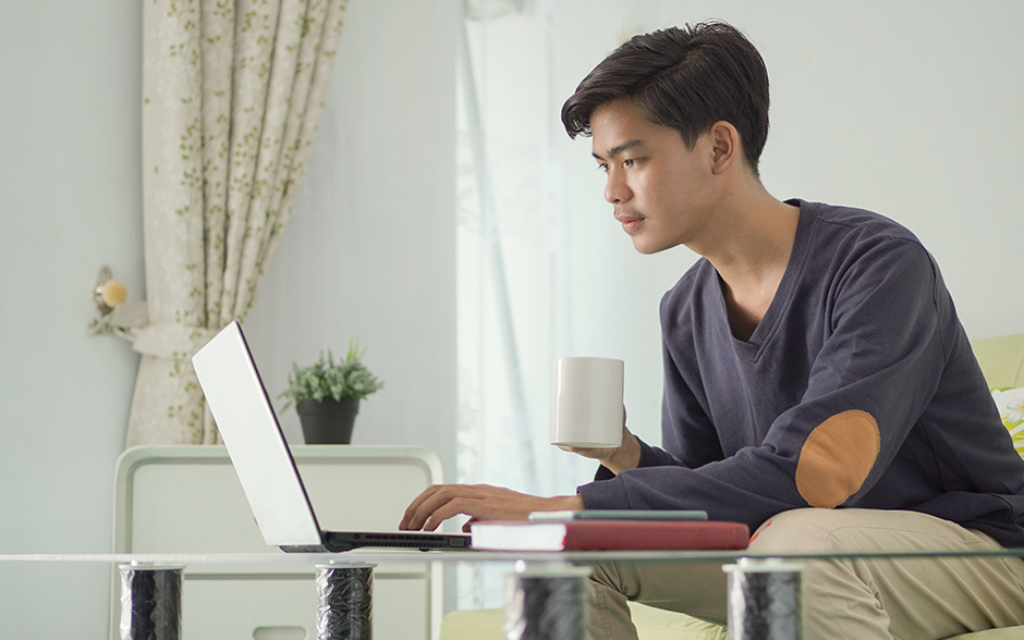 Young man working on a laptop at home while holding a coffee mug.