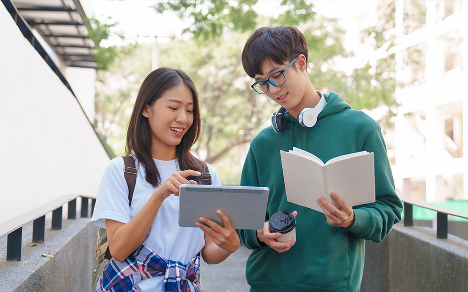 Two students discussing outdoors using a tablet and a notebook.