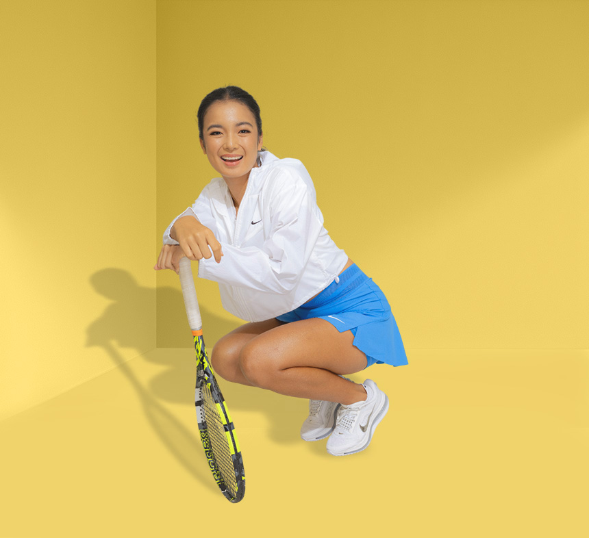 Alex Eala smiling in a white jacket and blue tennis skirt while holding a tennis racket.