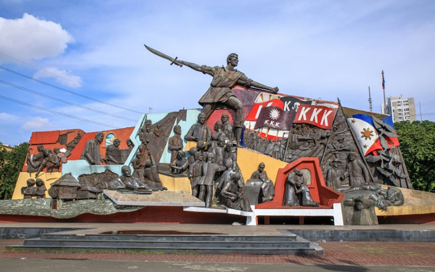 Monument with a central figure holding a sword, surrounded by historical reliefs and vibrant colors.